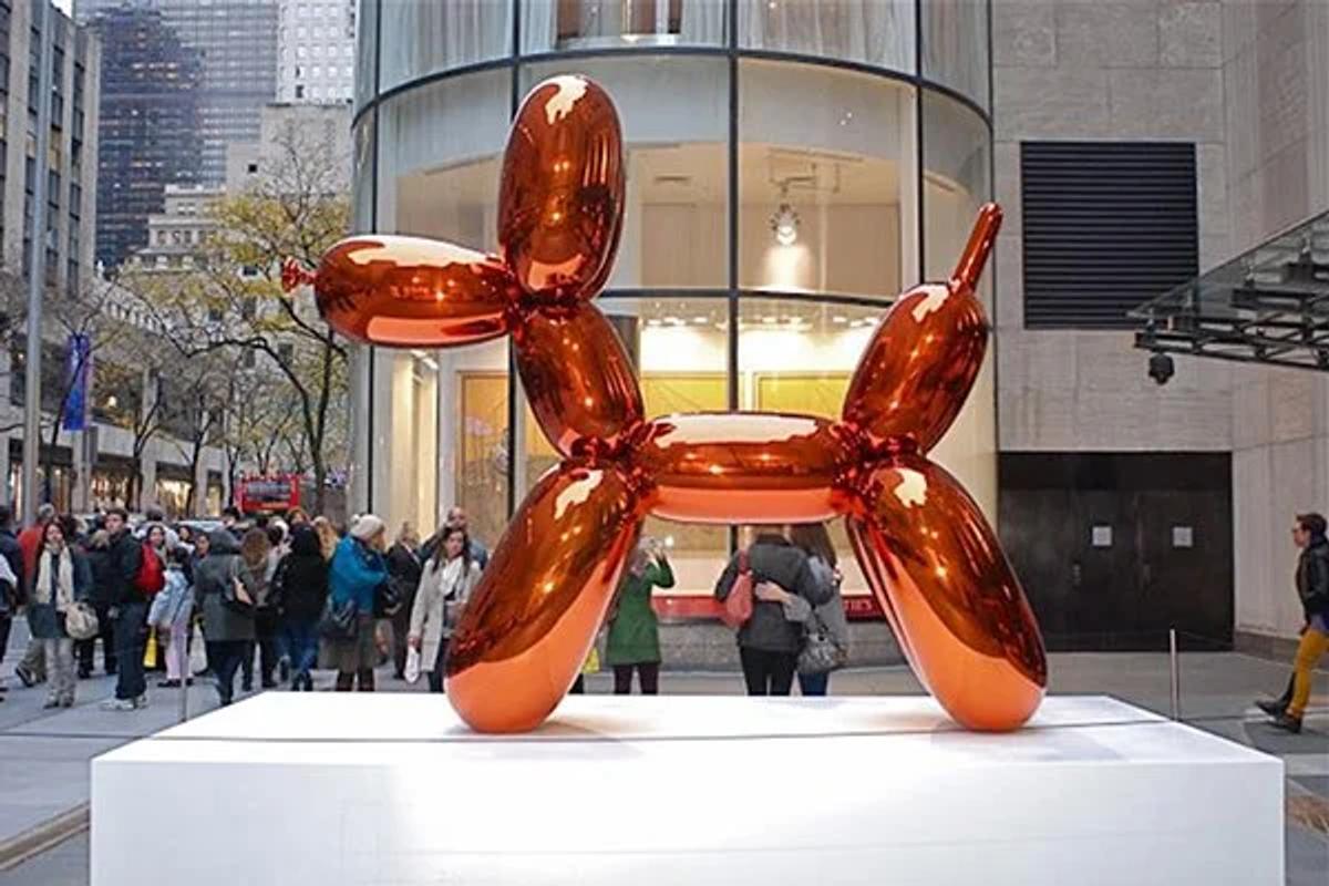 A large mirror polished orange balloon dog sculpture on a white plinth outside a curved glass building, with passers by behind it.