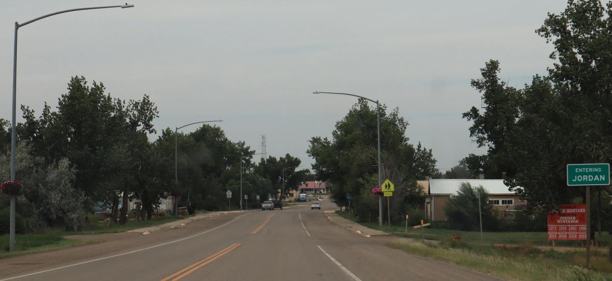 A two-lane highway running into the small town of Jordan, Montana, past cottonwoods and a green road sign reading ENTERING JORDAN.