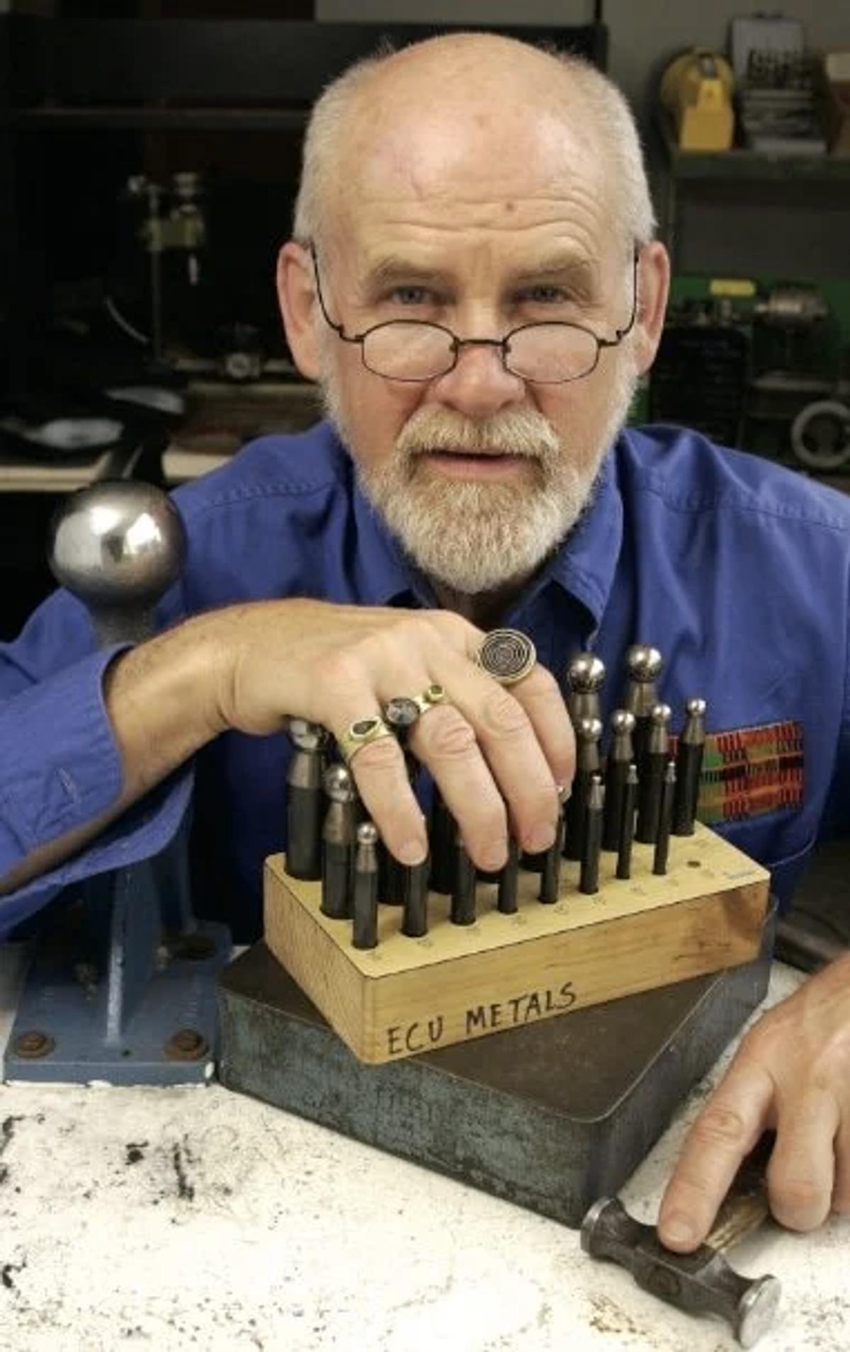 A silver haired man in glasses and a blue shirt at a metalworking bench, resting his hands beside a block of punches and a hammer.