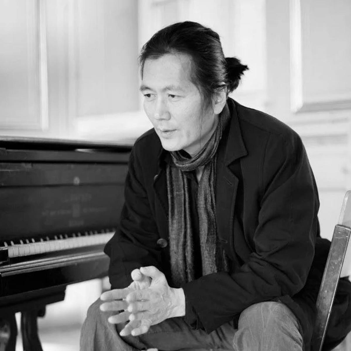 A black and white portrait of a man in a dark coat and scarf with his hair tied back, seated beside a grand piano.