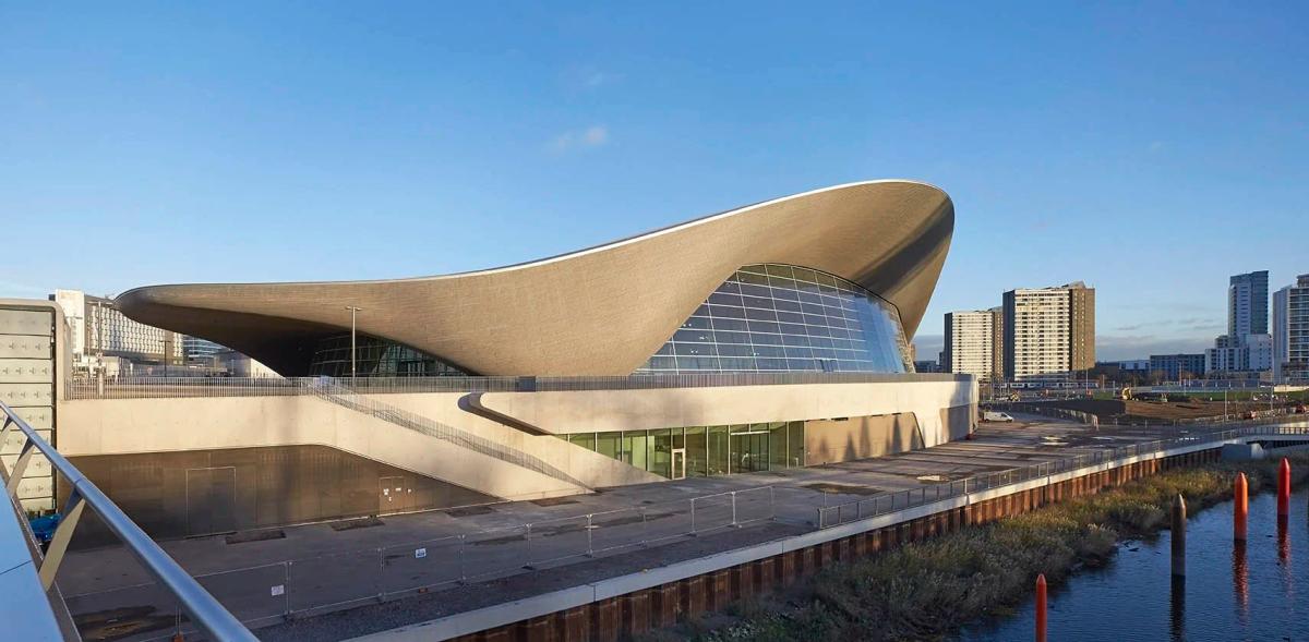 A low curved roofed aquatics building beside water, its long wave shaped roof sweeping out over a glazed facade, city towers behind.