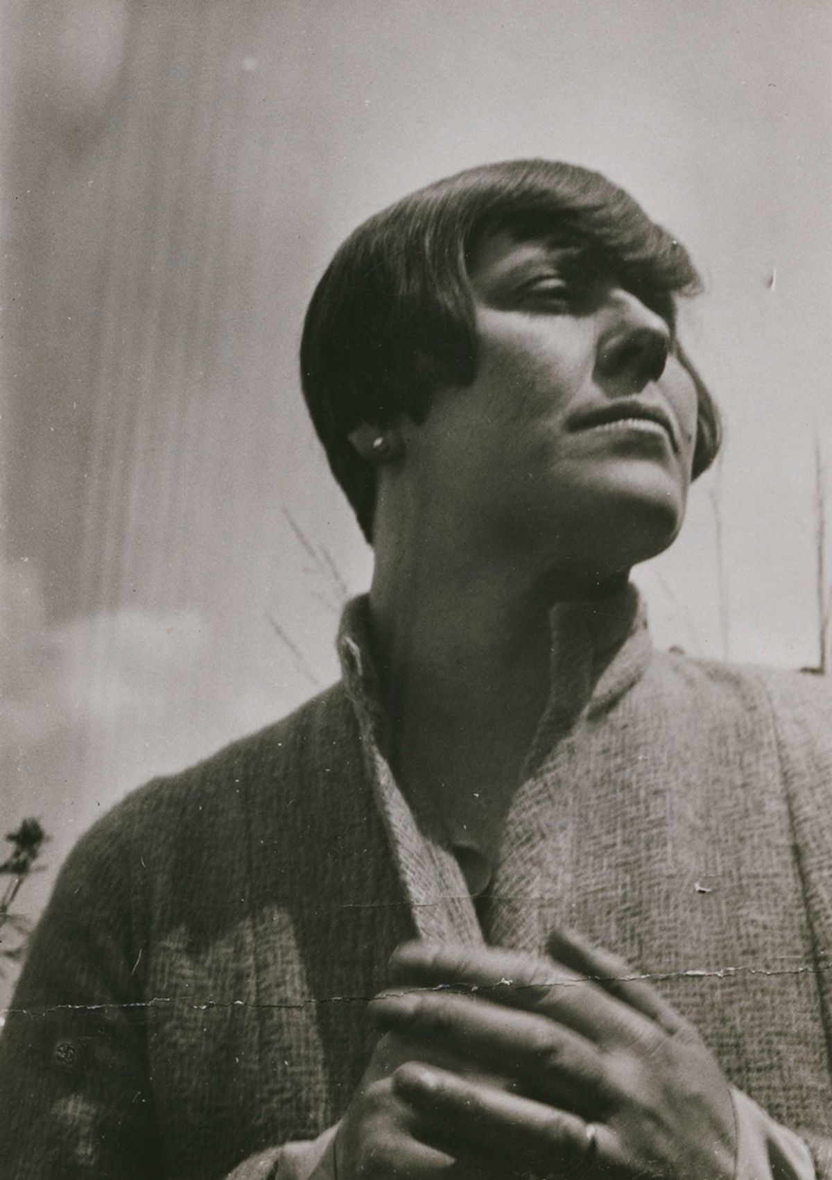A black and white portrait of a woman with a dark bob looking upward, hands clasped, in a textured jacket.