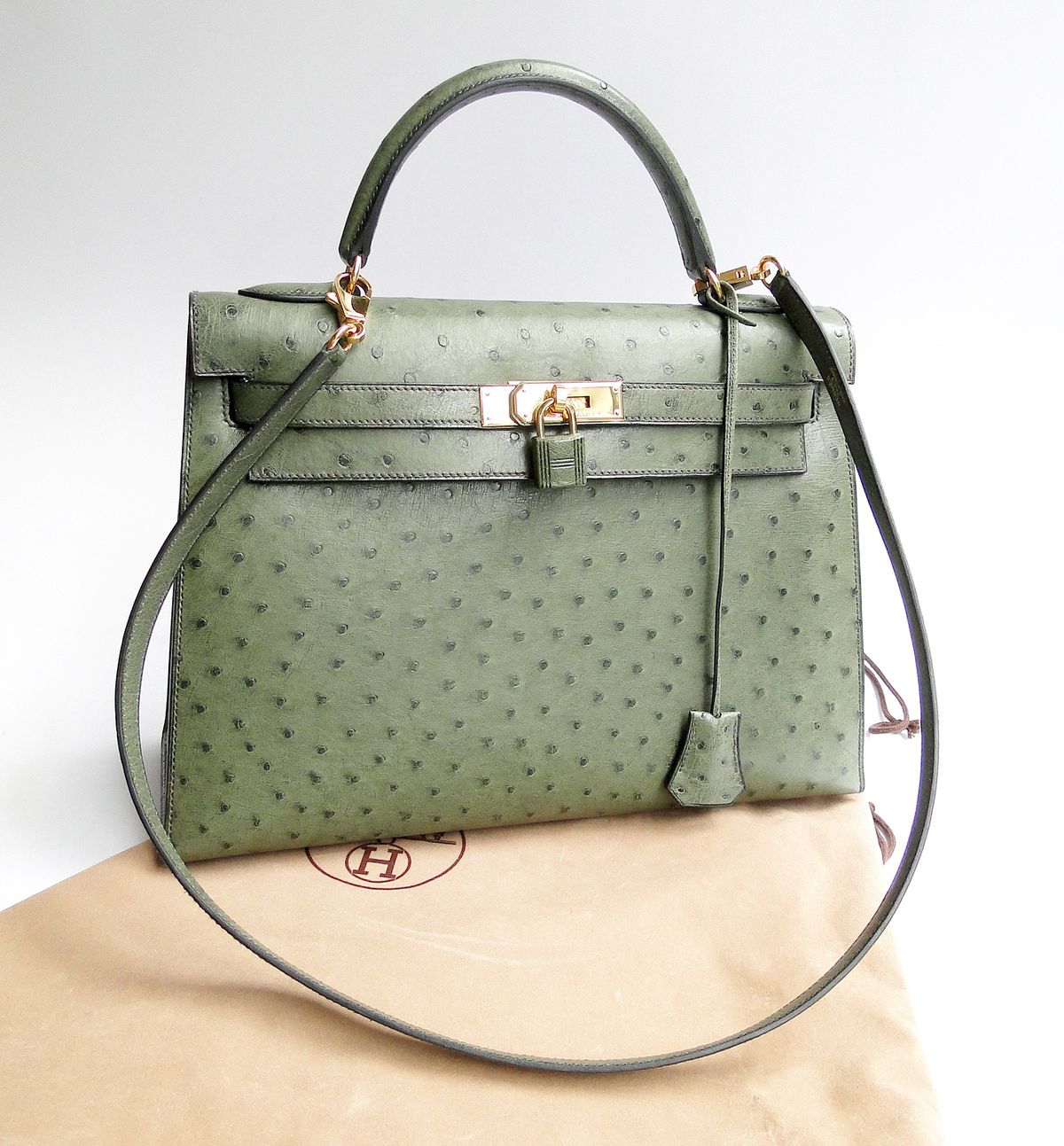 An olive green ostrich leather Hermes Kelly bag with gold hardware and a shoulder strap, standing on its dust bag.