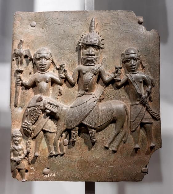 A cast bronze relief plaque showing a central mounted figure flanked by attendants, against a patterned ground, mounted on a museum stand with an accession number visible.