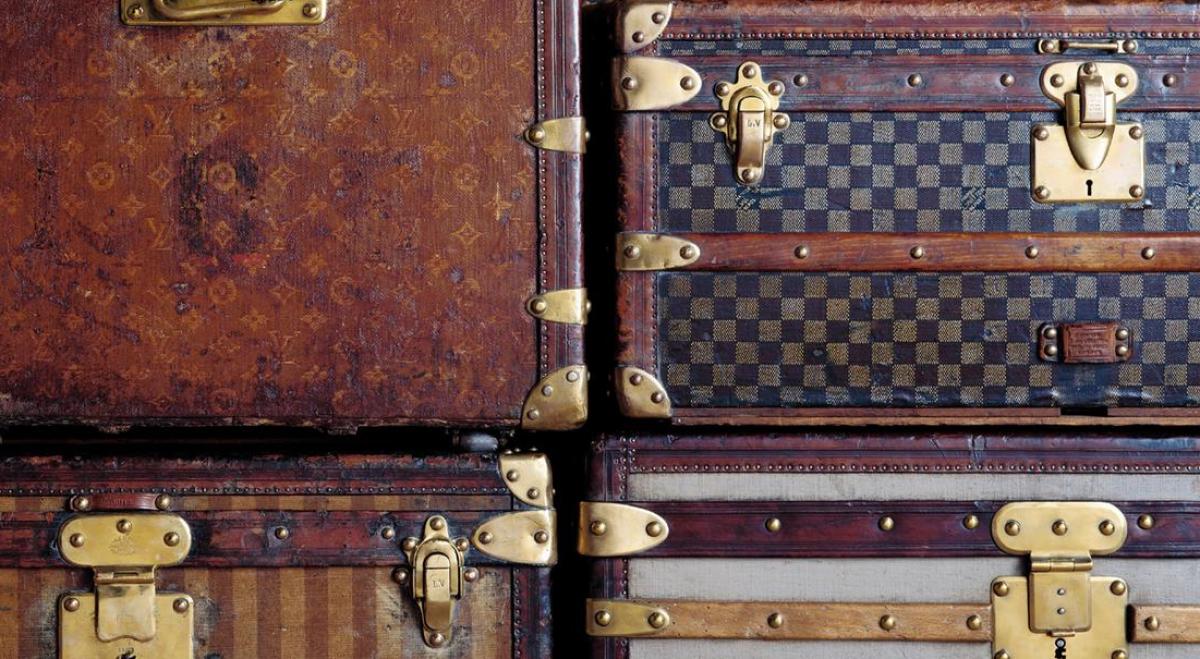 A stack of vintage travelling trunks in worn monogram and checkerboard canvas, bound with wooden battens and brass corners, locks and catches.