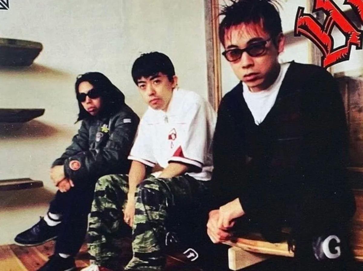 An archival photograph of three young men in dark glasses and streetwear seated on steps in front of a graffitied wall.