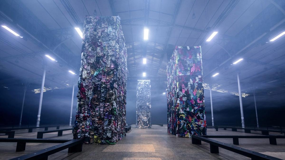 Two towering columns of compressed textile waste standing in a blue lit hall, benches and lighting rigs arranged around them.