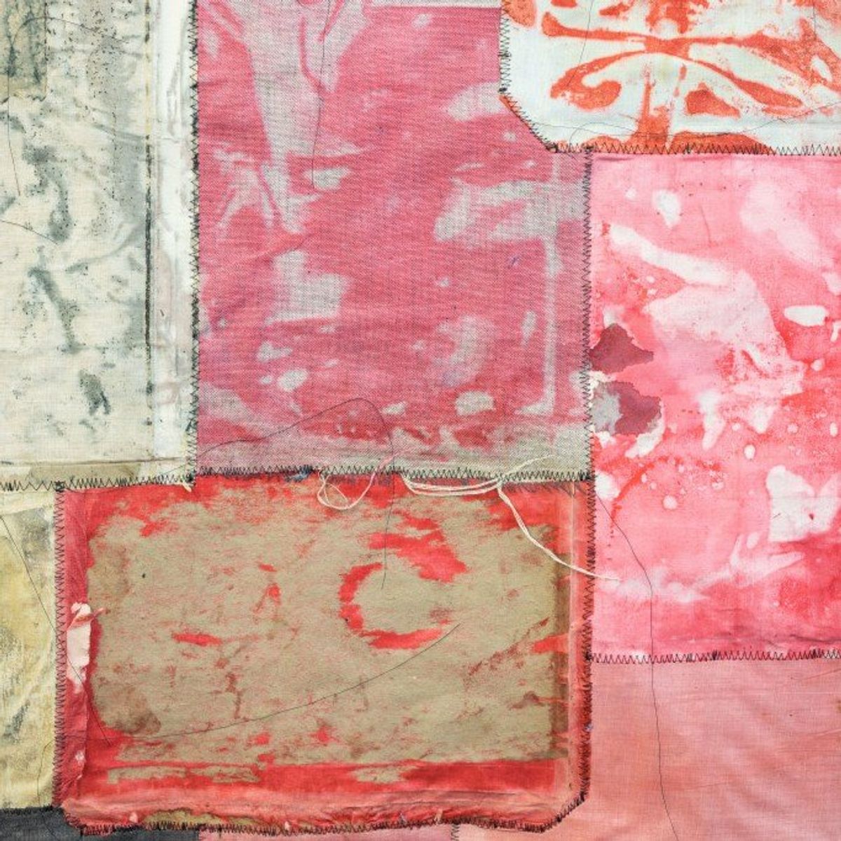 A close view of a pieced textile of stitched cloth rectangles in pink, cream and rust, with a faded red mark printed on the largest panel.