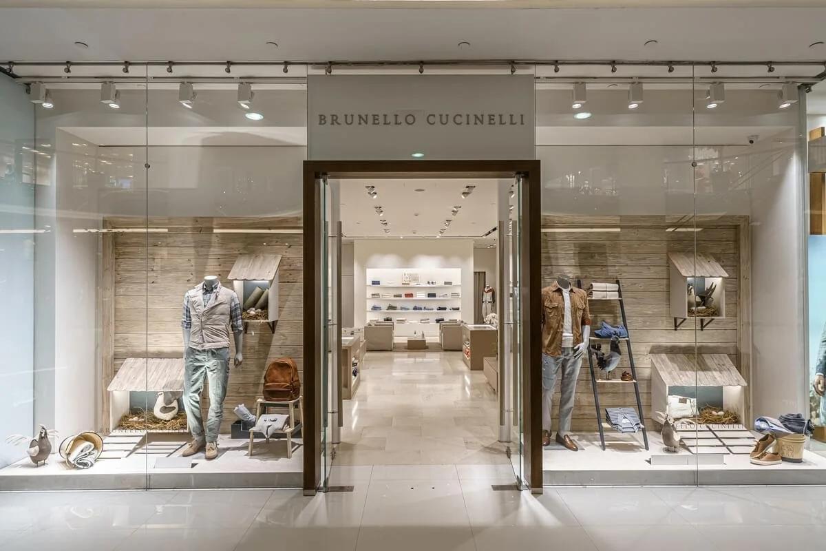 A pale stone shopfront with Brunello Cucinelli lettering above the entrance, dressed mannequins and knitwear displayed in the windows.
