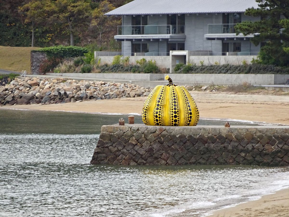 A large yellow pumpkin sculpture covered in black dots, standing alone at the end of a stone pier by grey sea.