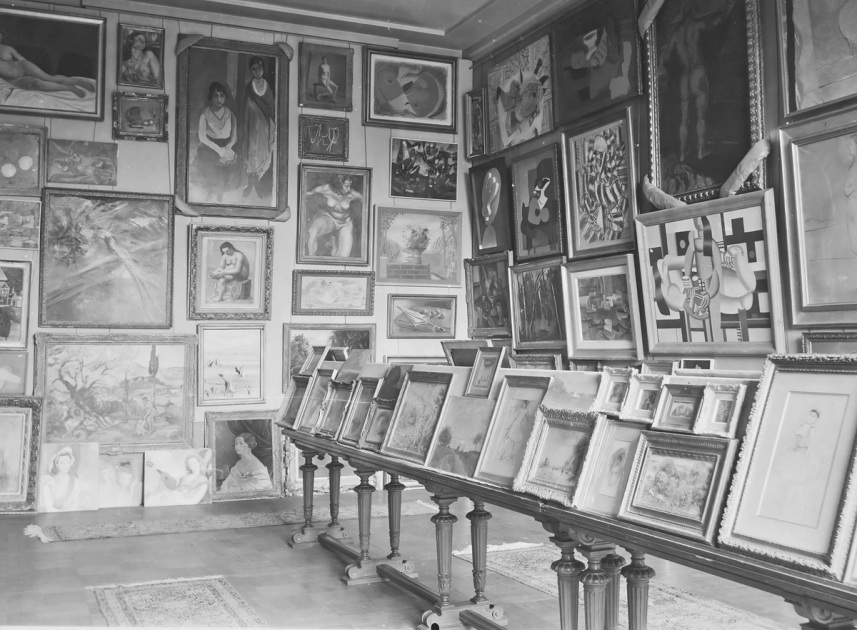 A black and white photograph of a gallery room packed floor to ceiling with framed paintings, more canvases propped in rows along a long table.