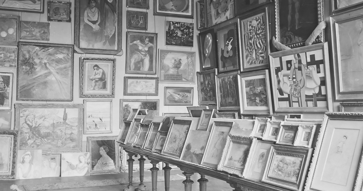 A black and white photograph of a gallery room packed floor to ceiling with framed paintings, more canvases propped in rows along a long table.