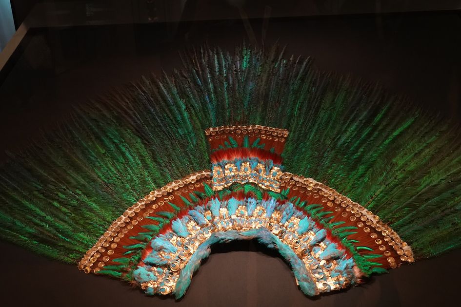 A wide semicircular fan of long green feathers above concentric bands of blue, red and brown feather mosaic set with small gold plates, displayed in a case.