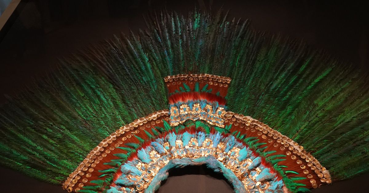 A wide semicircular fan of long green feathers above concentric bands of blue, red and brown feather mosaic set with small gold plates, displayed in a case.