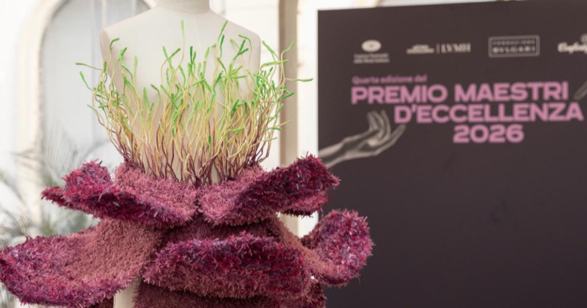 A dress form wearing a sculptural garment of deep pink felted ruffles topped with a collar of upright green grass, standing beside a dark board reading Premio Maestri d'Eccellenza 2026.