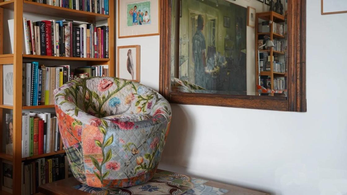 A corner of a book lined room with a floral upholstered armchair, a framed painting propped against the shelves and smaller pictures on the wall above.