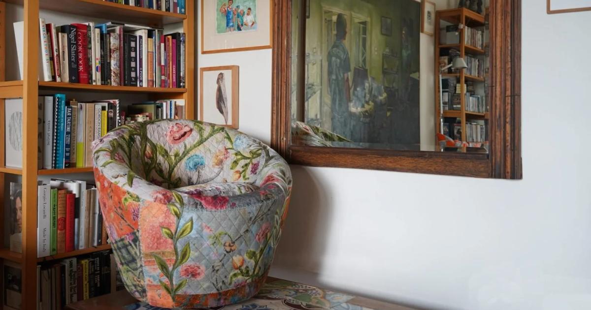 A corner of a book lined room with a floral upholstered armchair, a framed painting propped against the shelves and smaller pictures on the wall above.