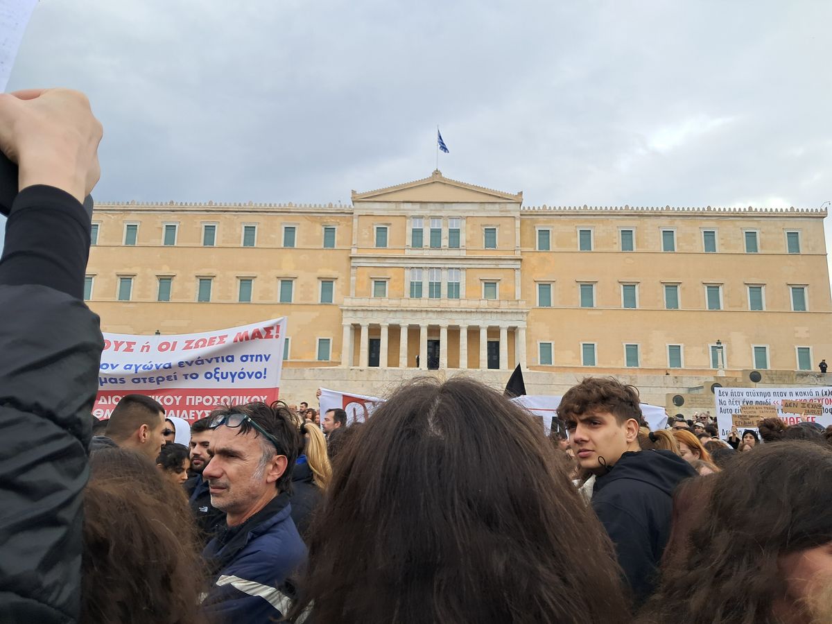 A crowd standing before the Hellenic Parliament in Athens under a heavy grey sky, holding banners and hand-written signs in Greek, with the Greek flag on the roof above.