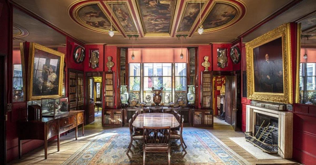 A red walled panelled room with a painted coffered ceiling, hung densely with gilt framed portraits, a long polished table and chairs on a patterned rug, a fireplace to one side.