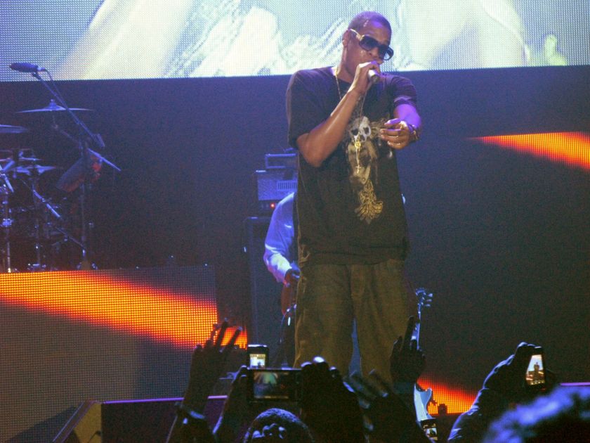 A performer in a dark t-shirt and gold chains stands at the front of a lit stage holding a microphone to his mouth, a large screen behind him and a drum kit to one side, with the raised hands of the crowd in the foreground holding up cameras and phones to record him.