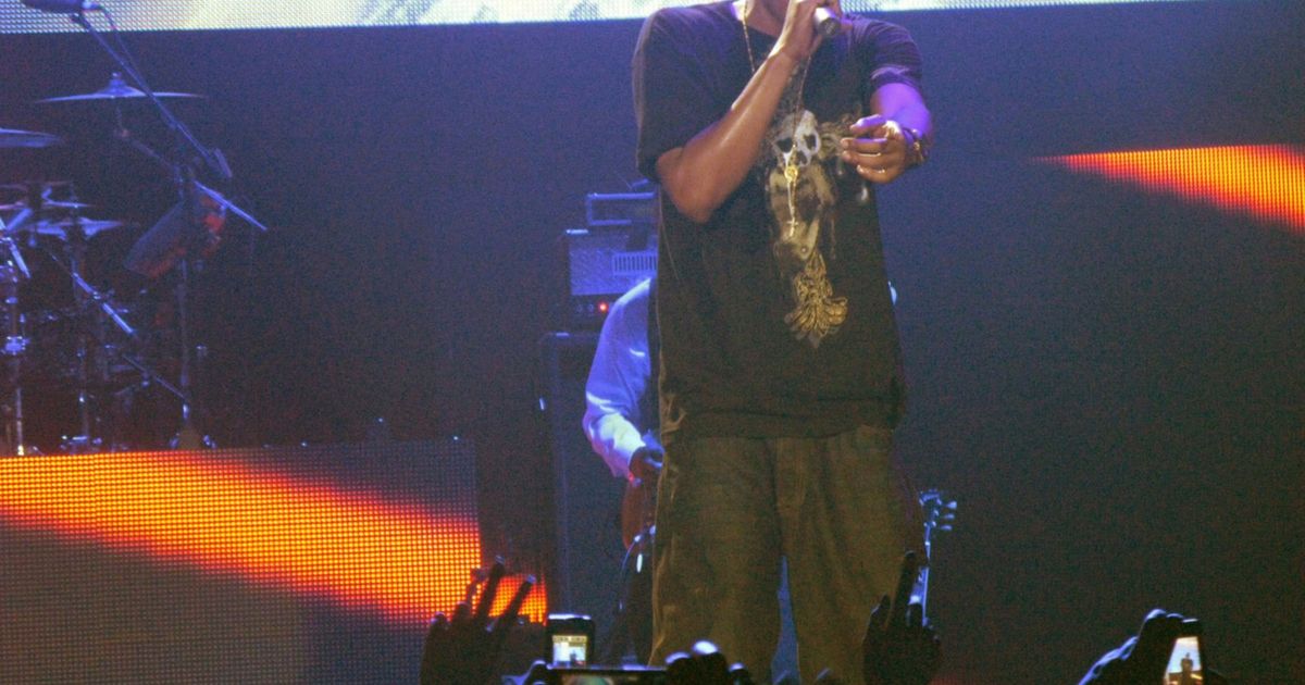 A performer in a dark t-shirt and gold chains stands at the front of a lit stage holding a microphone to his mouth, a large screen behind him and a drum kit to one side, with the raised hands of the crowd in the foreground holding up cameras and phones to record him.
