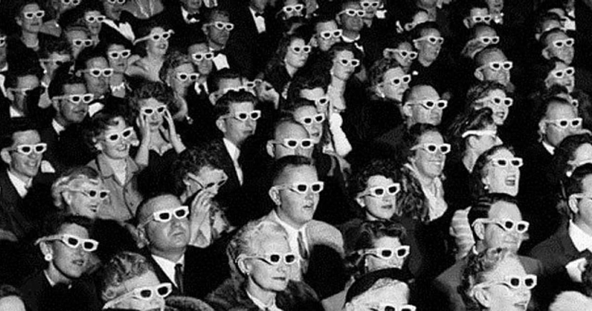 A black and white photograph of a packed cinema audience seated in rows, every person wearing paper 3D glasses and looking at the screen.
