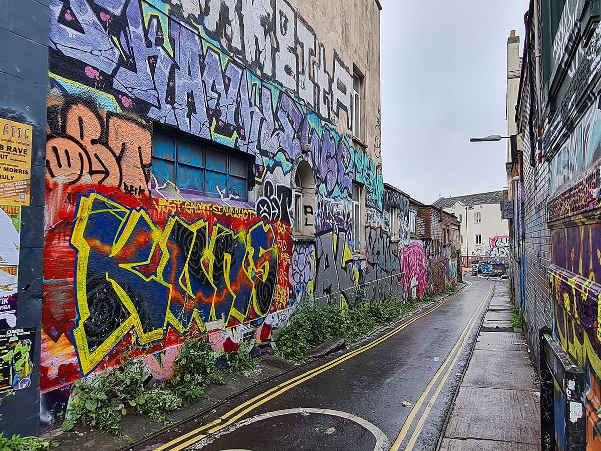 A narrow wet lane in Bristol with every wall surface covered in overlapping spray-painted work, receding towards a grey sky. No people are in the frame.
