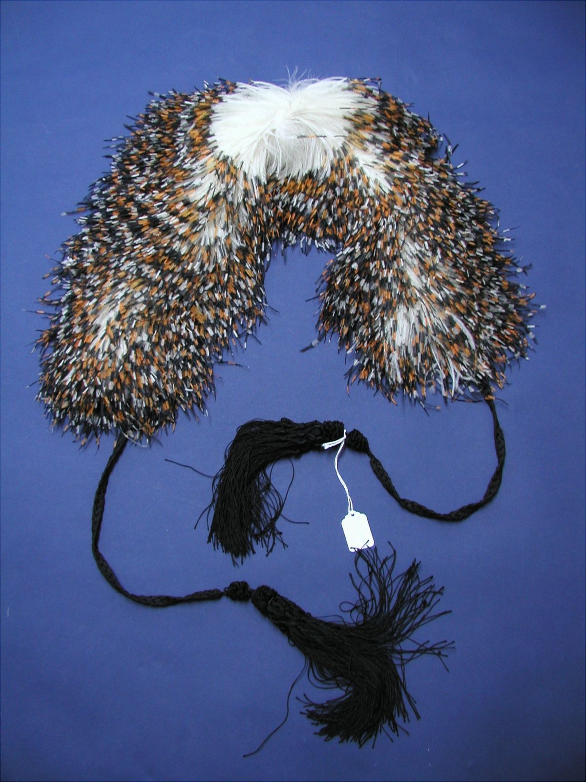A feather collar of brown, white and cream plumage arranged in dense overlapping rows, with long black tasselled cords hanging from each end, photographed against deep blue.