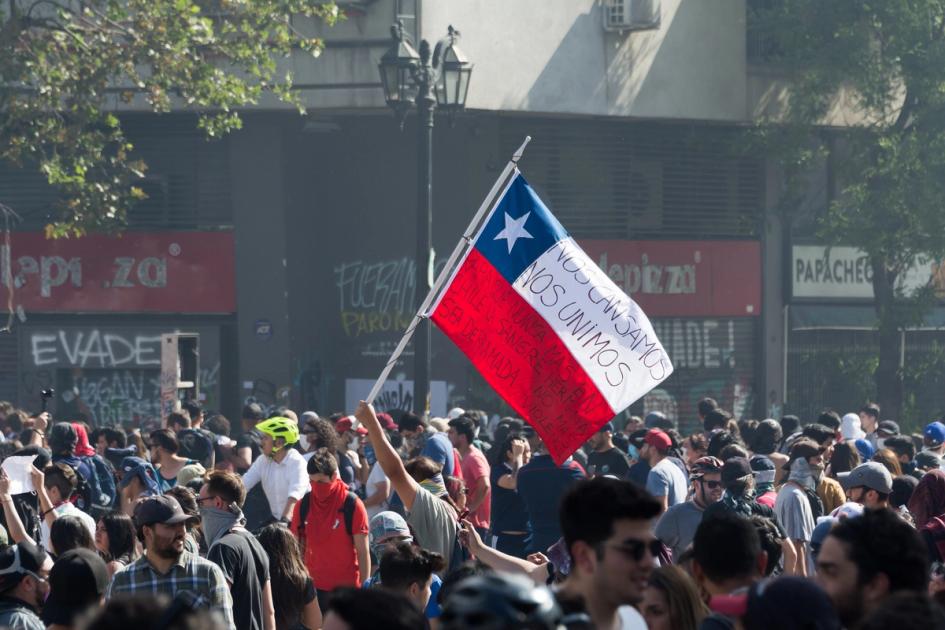 A Chilean flag raised above a dense crowd in a Santiago street, hand-lettered in black across its white and red bands. Behind the crowd, closed shop shutters are covered in graffiti, one word of which reads EVADE.