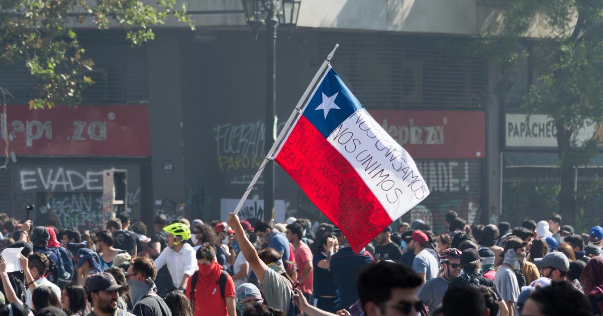 A Chilean flag raised above a dense crowd in a Santiago street, hand-lettered in black across its white and red bands. Behind the crowd, closed shop shutters are covered in graffiti, one word of which reads EVADE.