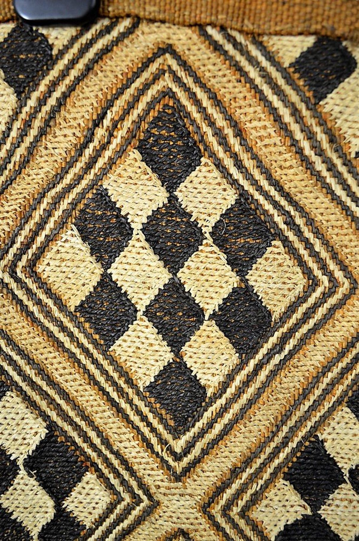 A close view of a woven raffia panel worked in concentric diamonds of black, cream and ochre with a dense stitched texture.