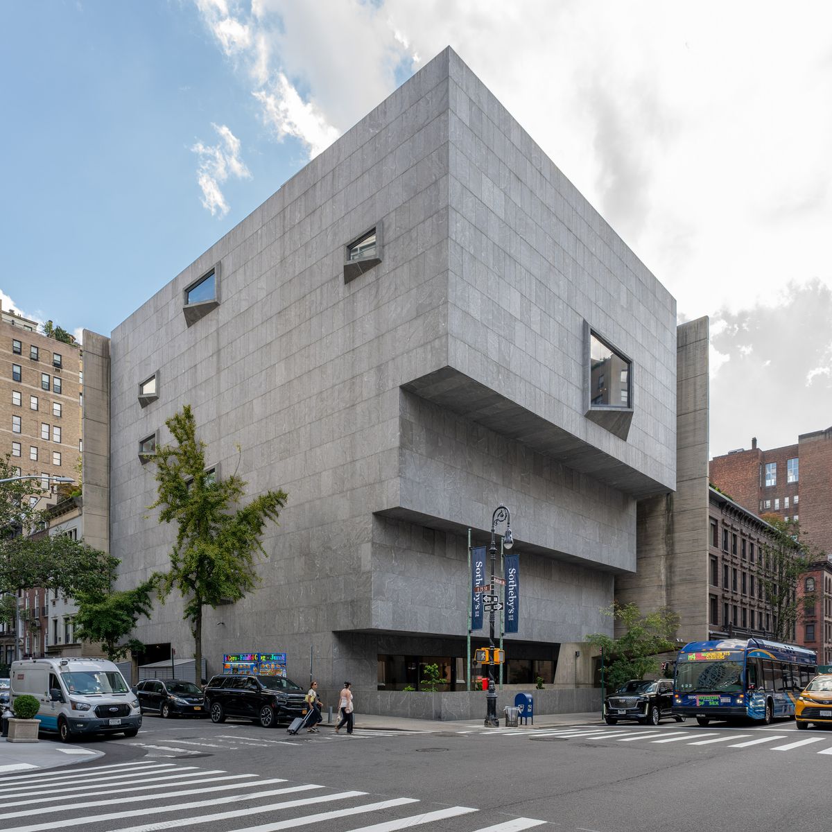 The grey granite Marcel Breuer building at 945 Madison Avenue seen from across the street, its upper floors cantilevered outward, with Sotheby's banners on the lamp posts.