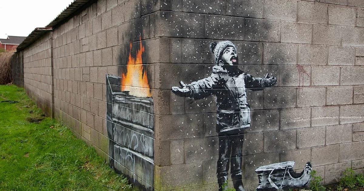 A stencilled mural on a grey breeze block garage: a child in hat and coat with arms outstretched catching what looks like snow, which is falling from a burning skip painted around the corner.