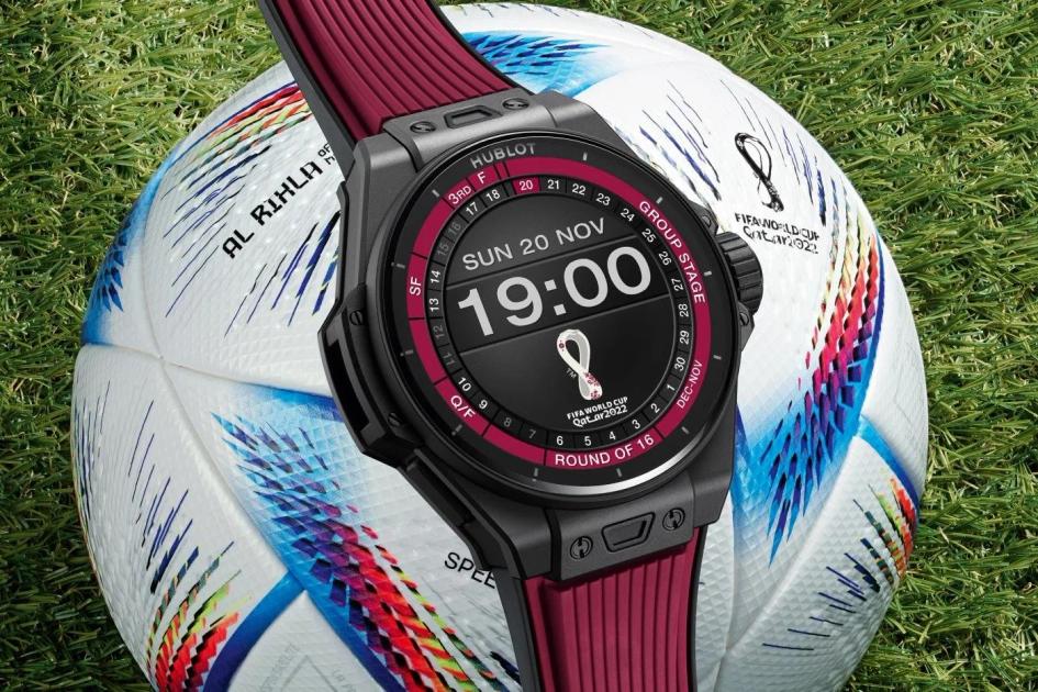A black and red sports watch resting against a white and blue World Cup football on grass, its face reading Sunday 20 November, 19:00.