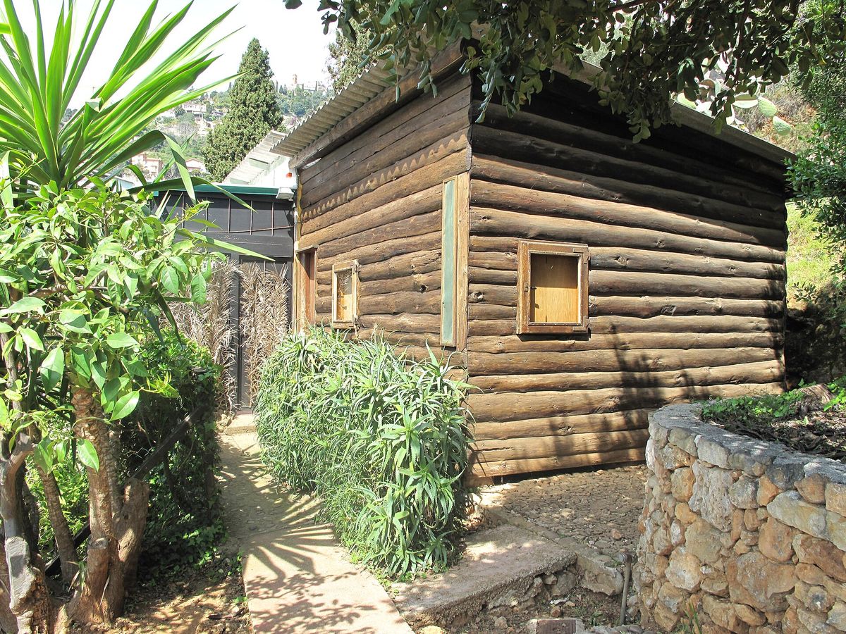 A small timber cabin with a shallow roof, standing among trees on a slope above the Mediterranean.