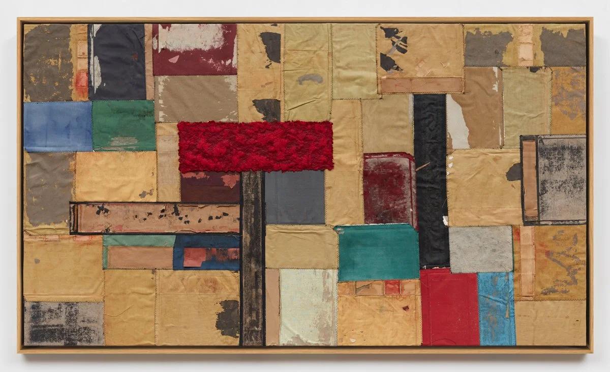 A framed panel assembled from salvaged paper and board pieces in ochre, red, blue and green, laid edge to edge in an irregular grid.