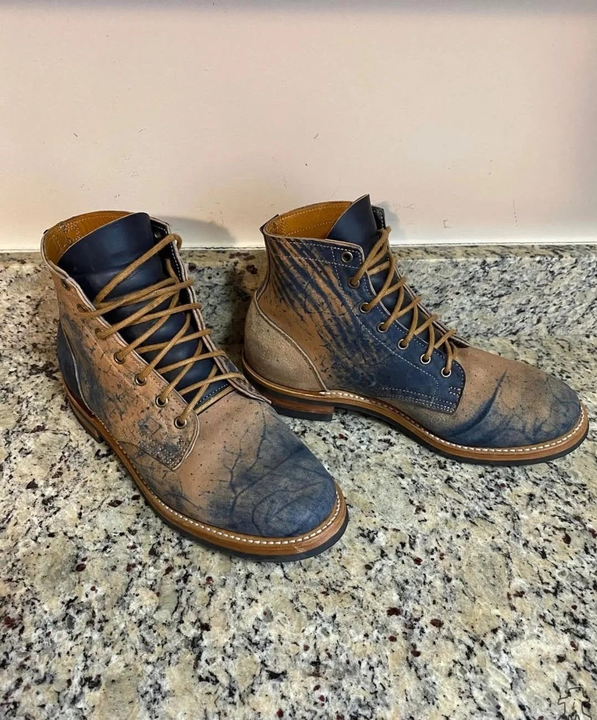 A pair of lace up leather boots on a granite floor, the leather dyed in mottled blue and brown with the pattern spreading unevenly across each shoe.
