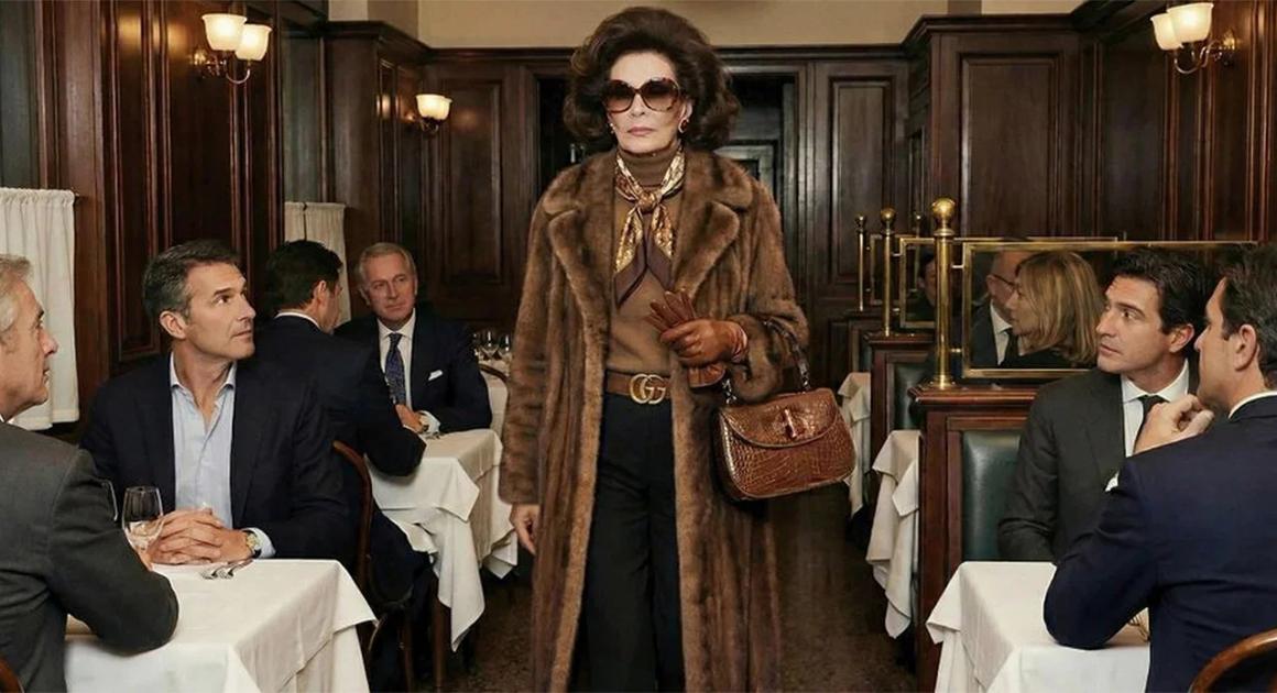 A woman in a fur coat, dark sunglasses and a monogrammed belt walking through a wood panelled restaurant, carrying a small crocodile handbag, past tables of seated men in suits.