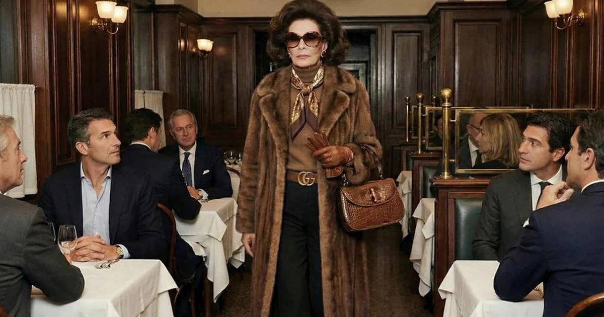 A woman in a fur coat, dark sunglasses and a monogrammed belt walking through a wood panelled restaurant, carrying a small crocodile handbag, past tables of seated men in suits.
