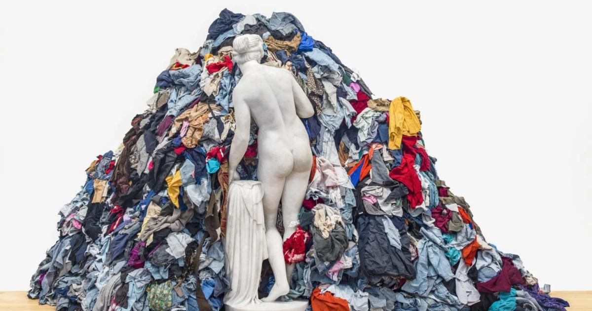 A classical white marble statue of a female nude standing partly buried in a large heaped mound of discarded clothing.