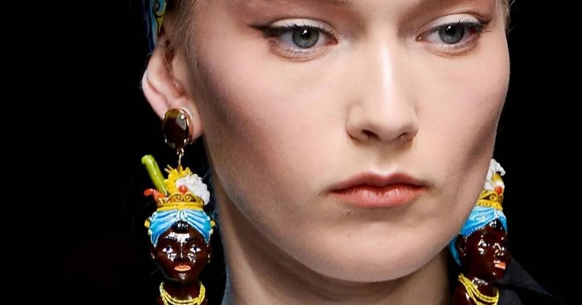 A close portrait of a model with pale skin and pulled back hair, wearing large ceramic drop earrings modelled as figures with dark faces in brightly coloured headwraps.