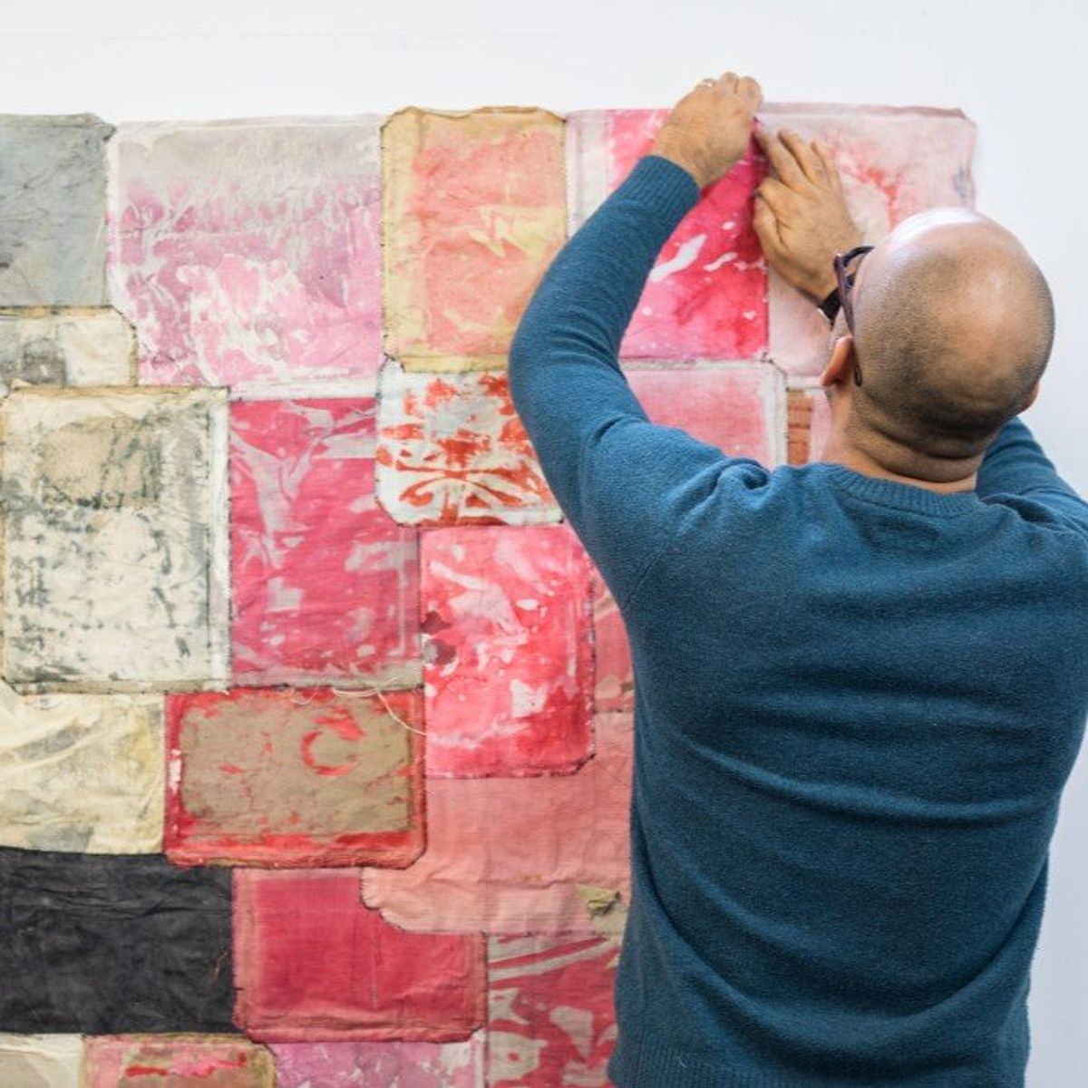 A man seen from behind reaching up to fix a large collaged work to a white wall, its surface a patchwork of pink, red, cream and black paper squares.