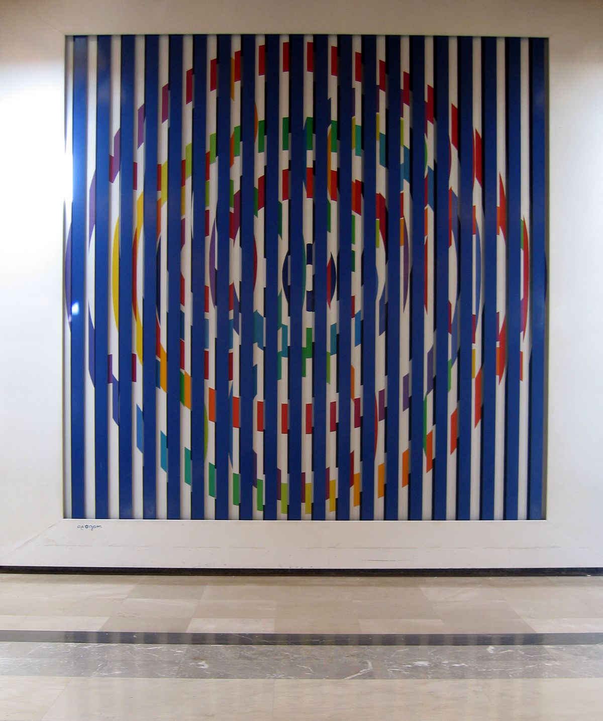 Large-scale kinetic panel work by Yaacov Agam in an institutional setting, showing his signature vertical ridge format with a circular spiral pattern in blue, red, green, and yellow