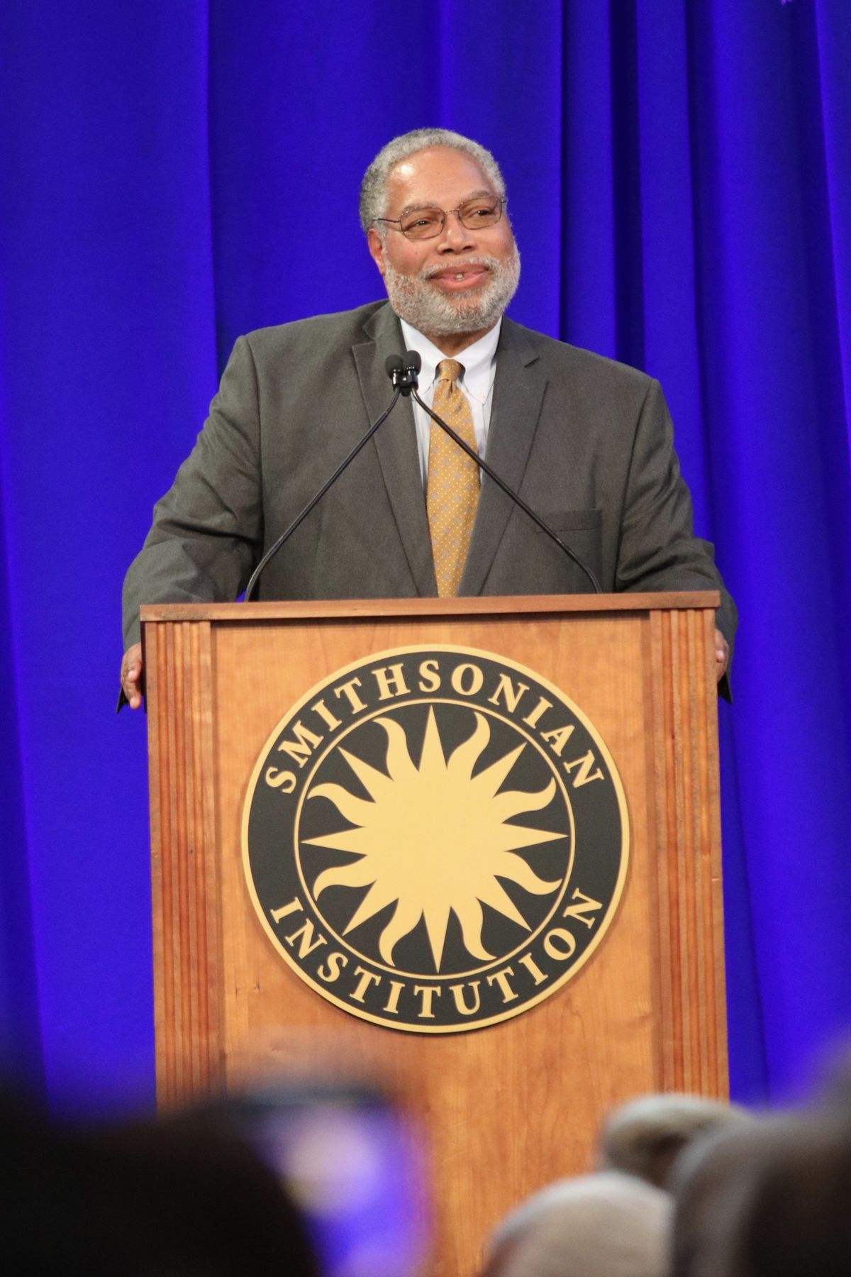 Lonnie G. Bunch III speaking at a wooden lectern bearing the Smithsonian Institution seal, against a blue curtain.