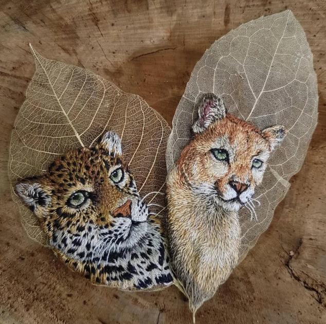 Two skeleton leaves laid on weathered wood, each painted with the head of a big cat, a spotted leopard on the left and a tawny cougar on the right.