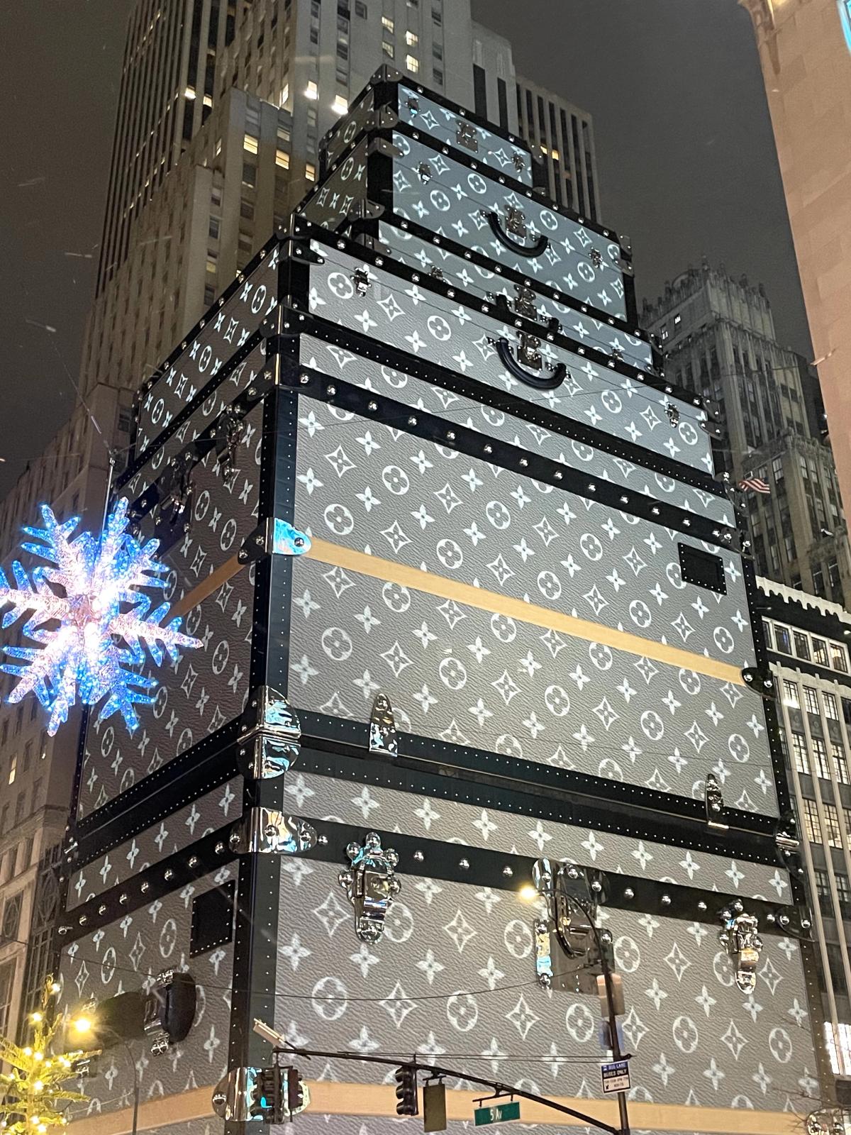 The same trunk-stack construction shed photographed at night in December, the monogrammed canvas lit from the street, with an illuminated snowflake ornament suspended at left.