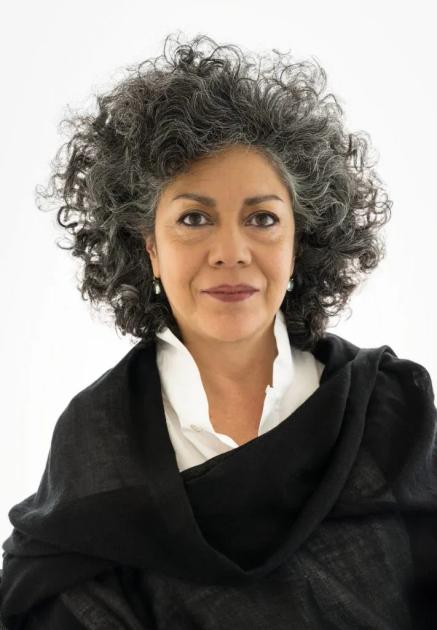 A portrait of a woman with grey curly hair against a white background, wearing a white shirt under a loosely draped black wrap.