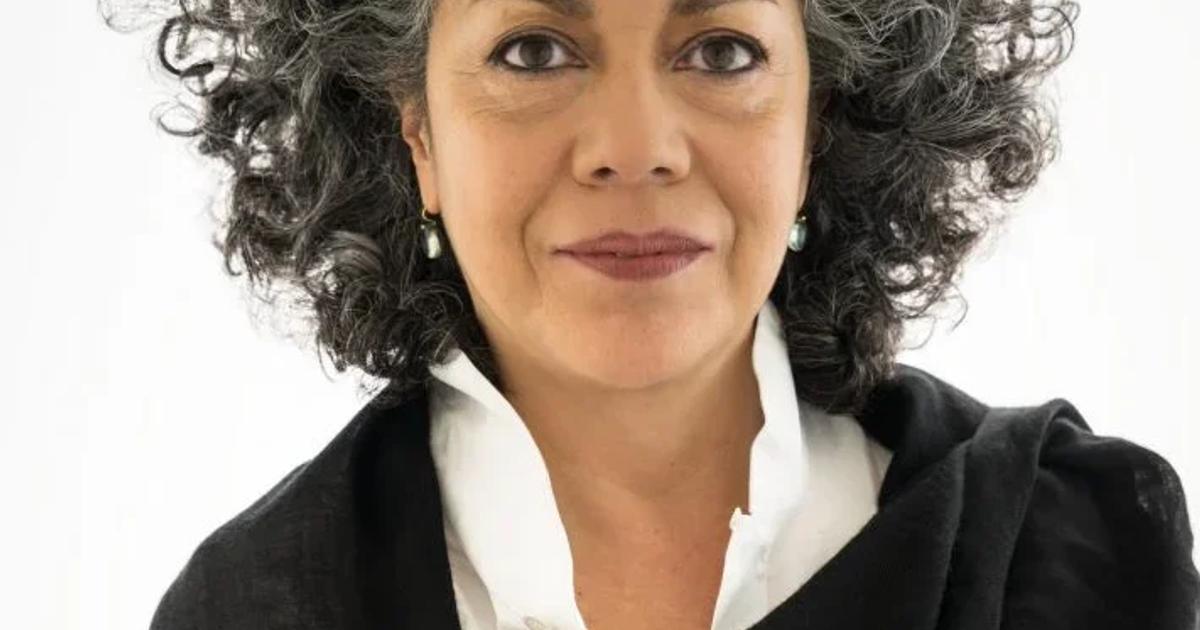A portrait of a woman with grey curly hair against a white background, wearing a white shirt under a loosely draped black wrap.
