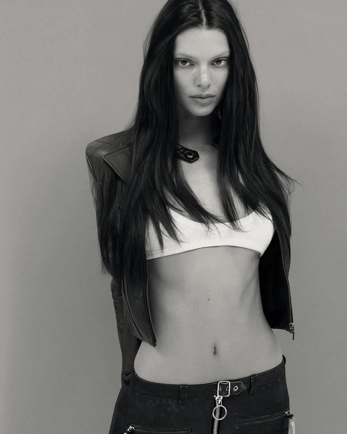 A black and white photograph of a woman in a cropped white top and leather jacket with long dark hair, standing against a plain wall.
