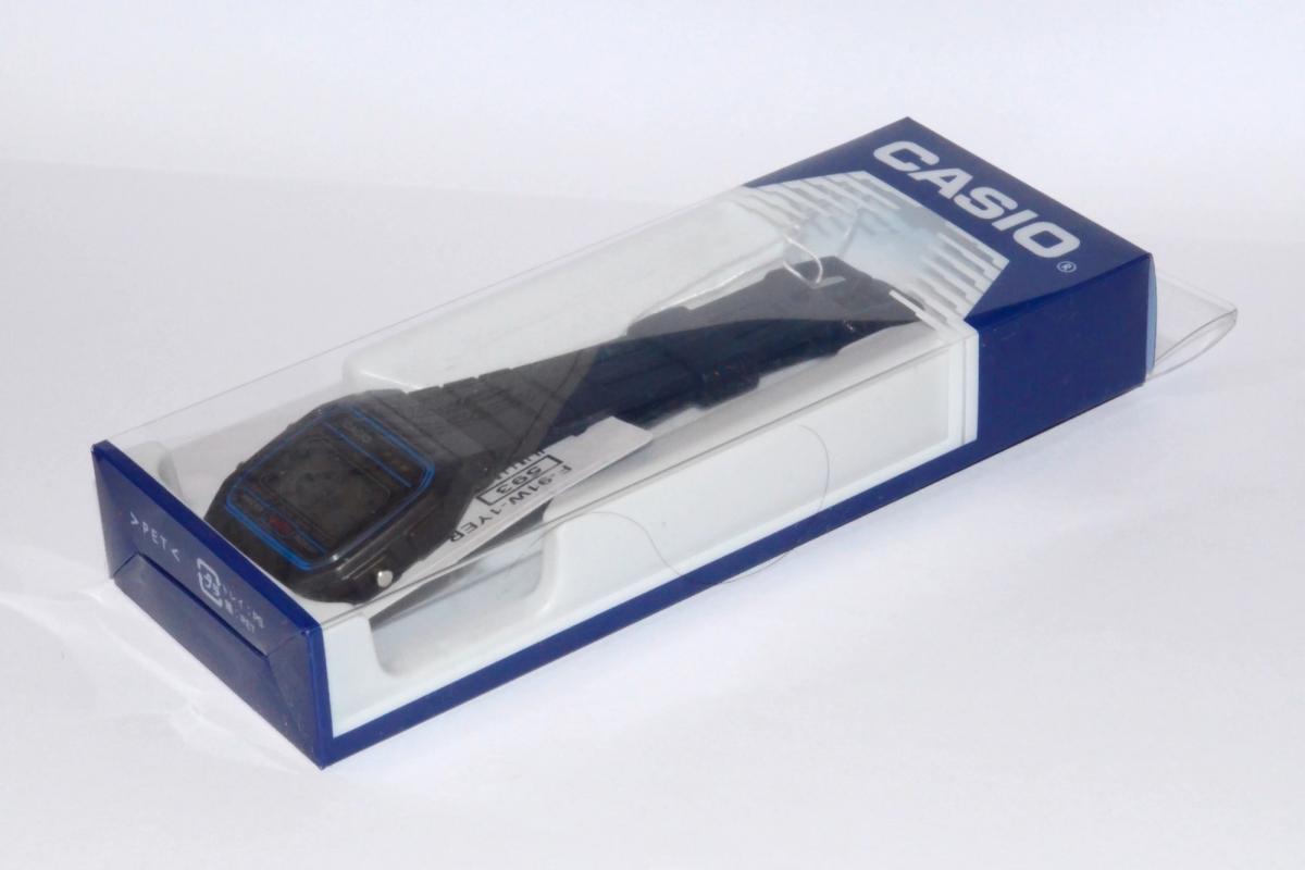 A Casio F-91W digital watch photographed beside its cardboard packaging, printed insert and paper tag.